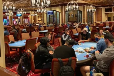 Etualalla pokeripöydän ympärillä istuu paljon pelaajia. Salissa on myös paljon muita pokeripöytiä. Lattia on väriltään punainen.