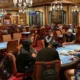 Etualalla pokeripöydän ympärillä istuu paljon pelaajia. Salissa on myös paljon muita pokeripöytiä. Lattia on väriltään punainen.