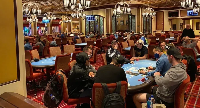 Etualalla pokeripöydän ympärillä istuu paljon pelaajia. Salissa on myös paljon muita pokeripöytiä. Lattia on väriltään punainen.