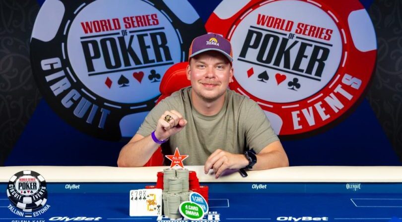Mies istuu pokeripöydässä. Mies hymyilee ja hänellä on päässään lippis. Hänellä on toinen käsinyrkissä ja yhdessä sormessa on sormus.