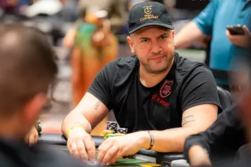 Mies istuu pokeripöydässä. Hänellä on päällään musta t-paita ja musta lippis. Miehen kädet on vihreällä pöydällä.