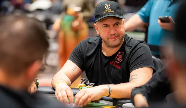 Mies istuu pokeripöydässä. Hänellä on päällään musta t-paita ja musta lippis. Miehen kädet on vihreällä pöydällä.