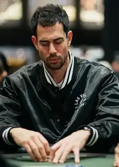 Mies istuu pokeripöydässä. Hänellä on päällään musta pilottitakki. Vieressä istuu toinen mies, jolla on päässään huppu ja lippis.