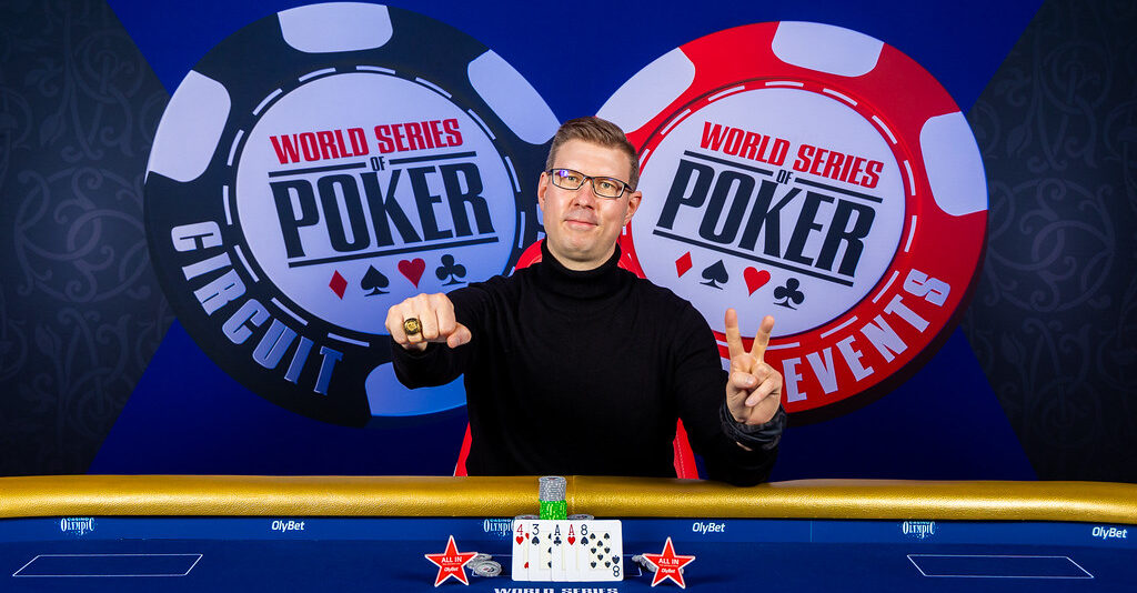 Mies istuu pokeripöydässä. Toisessa kädessä miehellä on kaksi sormea pystyssä ja toinen käsi on nyrkissä. Pöydällä on pelikortteja.