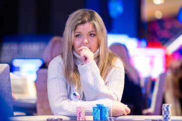 Vaaleahiuksinen nainen istuu pöydän ääressä vaaleassa paidassa, hän on peittänyt suunsa kädellään. Taustalla näkyy mustaa ja sinistä.