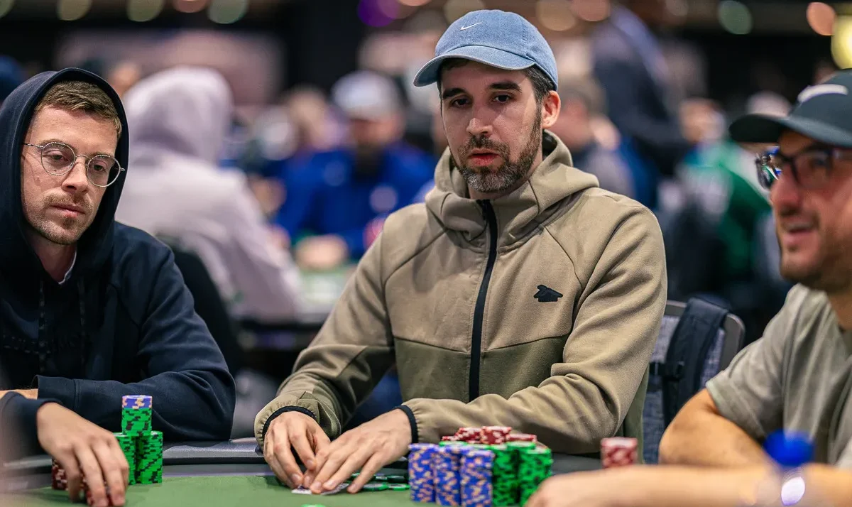 Miehet istuu pokeripöydässä. Heillä on edessään pelimerkkejä. Kahdella miehistä on päässään lippikset. Takana näkyy lisää ihmisiä.