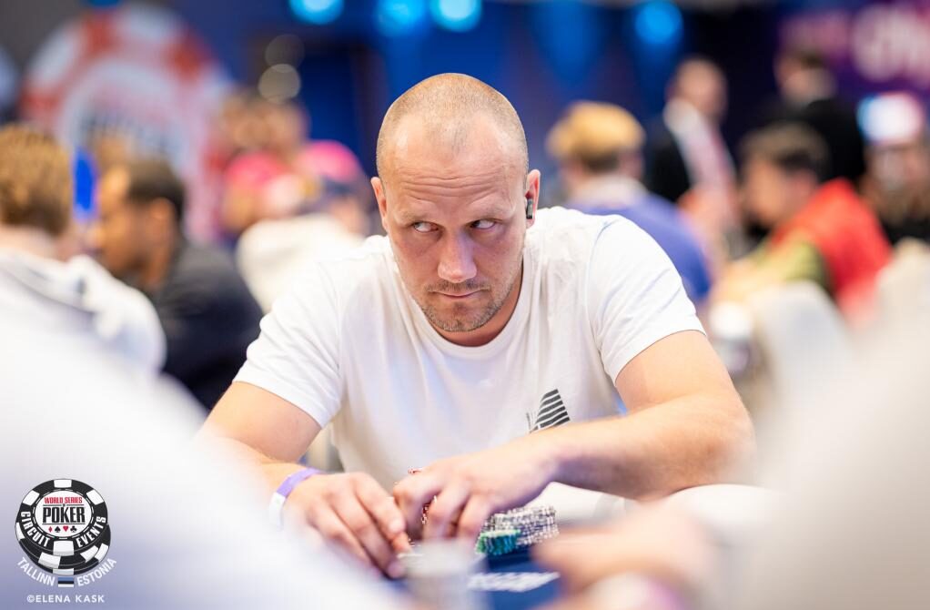 Kalju mies istuu pokeripöydässä. Hänellä on päällään valkea t-paita. Hänellä on otsa hieman kurtussa ja hän katsoo sivulle.