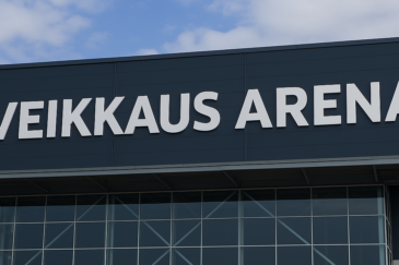 Kuvassa näkyy rakennusken seinää. Seinässä lukee mustalla pohjalla Veikkaus Arena. Taustalla näkyy sinivalkoinen taivas.