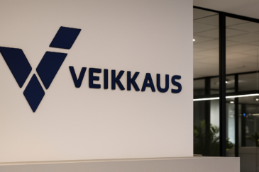 Valkoisella seinällä on sininen Veikkauksen logo ja lukee Veikkaus. Seinän vieressä on ikkunoita, jotka on rajattu karmeilla.
