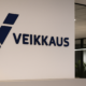 Valkoisella seinällä on sininen Veikkauksen logo ja lukee Veikkaus. Seinän vieressä on ikkunoita, jotka on rajattu karmeilla.