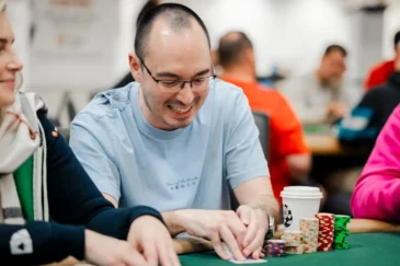 Mies istuu pokeripöydässä. Hän katsoo korttejaan ja nauraa samalla. Mies on lähes kalju ja hänellä on silmälasit päässään.