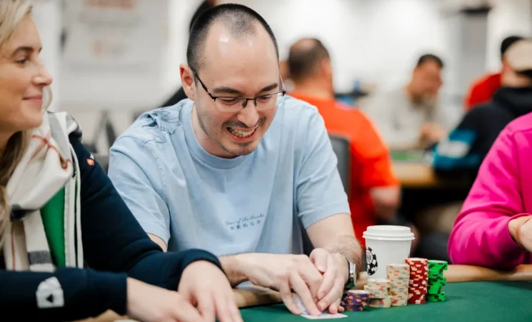 Mies istuu pokeripöydässä. Hän katsoo korttejaan ja nauraa samalla. Mies on lähes kalju ja hänellä on silmälasit päässään.