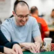 Mies istuu pokeripöydässä. Hän katsoo korttejaan ja nauraa samalla. Mies on lähes kalju ja hänellä on silmälasit päässään.