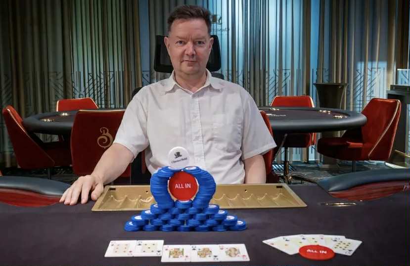 Mies istuu pokeripöydässä. Miehellä on päällään valkoinen kauluspaita. Pöydällä on pelimerkkejä sekä pelimerkeistä kasattu teos.