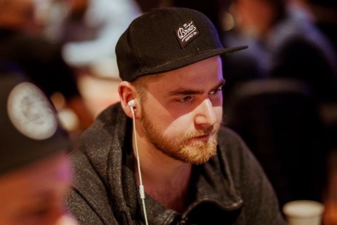 Miehellä on valkoiset kuulokkeet korvilla ja musta lippis päässä. Hän istuu pokeripöydän ääressä vakavalla naamalla.