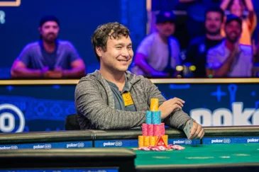 Mies istuu pokeripöydässä ja hymyilee. Hänen edessään pöydällä on erivärisiä pelimerkkejä torneissa. Takana näkyy ihmisiä.