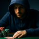Mies istuu pokeripöydässä. Hänellä on sininen huppu päässään. Miehen käden alla on kaksi pelikorttia ja vieressä pelimerkkejä.