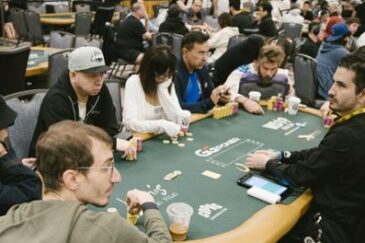 Pokeripöydässä istuu kahdeksan pelaaja ja jakaja. Taustalla näkyy lisää vihreäverkaisia pokeripöytiä sekä ihmisiä pöydissä.
