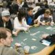 Pokeripöydässä istuu kahdeksan pelaaja ja jakaja. Taustalla näkyy lisää vihreäverkaisia pokeripöytiä sekä ihmisiä pöydissä.