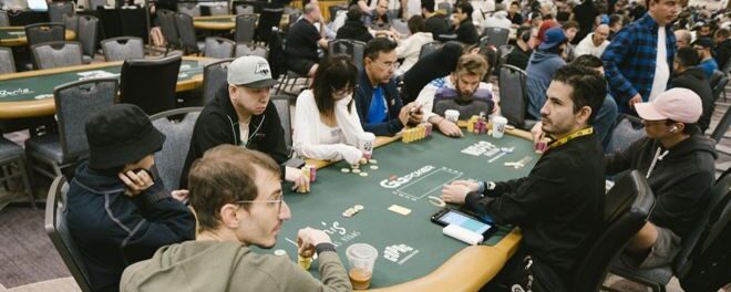 Pokeripöydässä istuu kahdeksan pelaaja ja jakaja. Taustalla näkyy lisää vihreäverkaisia pokeripöytiä sekä ihmisiä pöydissä.
