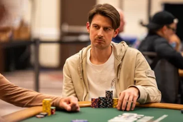 Mies istuu pokeripöydässä. Hänellä on päällään vaalea huppari, joka on auki. Hupparin alla miehellä on valkoinen t-paita.