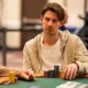 Mies istuu pokeripöydässä. Hänellä on päällään vaalea huppari, joka on auki. Hupparin alla miehellä on valkoinen t-paita.