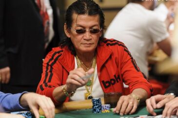 Scotty Nguyen istuu pokeripöydän ääressä punaisessa takissa ja aurinkolasit päässään edessään merkkipino
