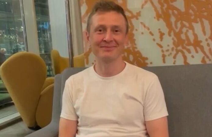 Antti Hakala valkoisessa T-paidassa istuu ruskeavalkoisen seinän edessä urheilukello kädessään.