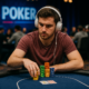 Mies istuu pokeripöydän ääressä harmaat kuulokkeet päässään ja punertavassa paidassa. Edessään hänellä on pokerimerkkejä ja kortit.
