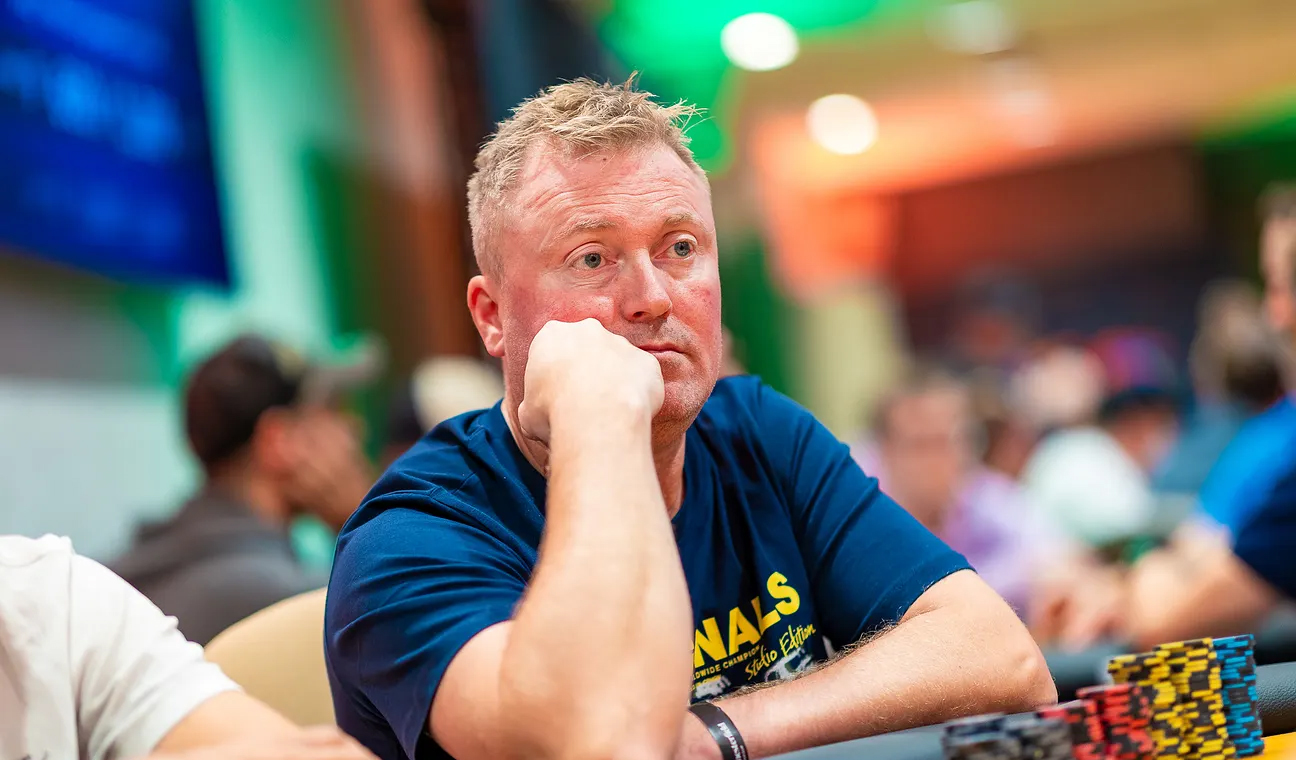 Mies nojaa nyrkkiinä pokeripöydässä istuen. Hänen ilmeensä on poissaoleva ja pohdiskeleva. Takana näkyy värikkäitä valoja.