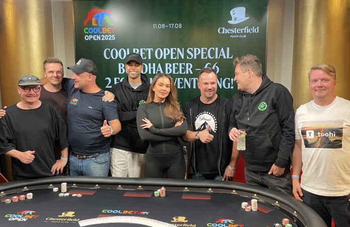 Pokeripöydän takana seisoo kahdeksan ihmistä, jotka hymyilevät ja halailevat toisiaan. Pöydän takana on kultaiset verhot.