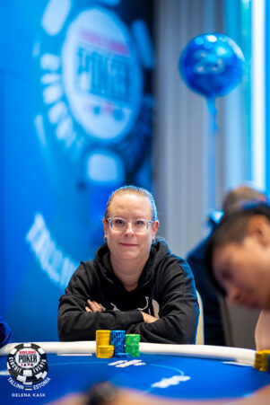 Nainen, jolla on silmälasit istuu pokeripöydän ääressä mustassa hupparissa. Tausta on sininen ja valkoinen, etualalla näkyy mies.