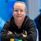 Nainen, jolla on silmälasit istuu pokeripöydän ääressä mustassa hupparissa. Tausta on sininen ja valkoinen, etualalla näkyy mies.