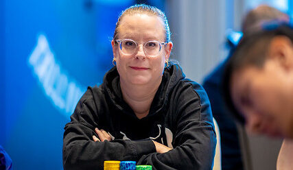 Nainen, jolla on silmälasit istuu pokeripöydän ääressä mustassa hupparissa. Tausta on sininen ja valkoinen, etualalla näkyy mies.