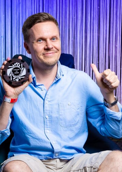 Mies pitää kädessään pokaalia ja näyttää peukkua. Hänellä on päällään sininen kauluspaita.