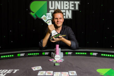 Mies istuu pokeripöydässä. Hänellä on päällään musta kauluspaita ja käsissään pokaali. Miehen edessä pöydällä on pelimerkkejä.