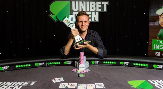 Mies istuu pokeripöydässä. Hänellä on päällään musta kauluspaita ja käsissään pokaali. Miehen edessä pöydällä on pelimerkkejä.