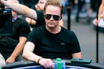 Mies istuu pokeripöydässä. Hänellä on päällään musta t-paita ja mustat aurinkolasit. Miehen edessä pöydällä on pelimerkkejä.