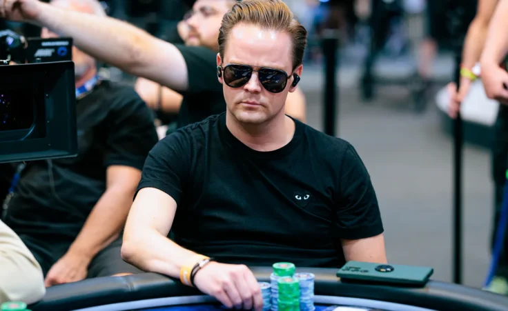 Mies istuu pokeripöydässä. Hänellä on päällään musta t-paita ja mustat aurinkolasit. Miehen edessä pöydällä on pelimerkkejä.
