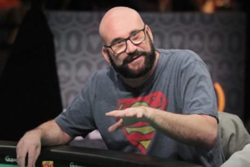 Mies istuu pokeripöydän ääressä, hänellä on silmälasit, musta parta ja harmaa paita. Hän tekee elettä kädellään. tausta on tumma.