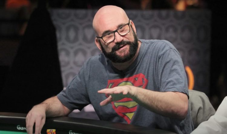 Mies istuu pokeripöydän ääressä, hänellä on silmälasit, musta parta ja harmaa paita. Hän tekee elettä kädellään. tausta on tumma.