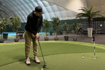 Mies mustissa vaatteissa on sisähallissa puttaamassa golf-mailalla golf-palloa. Alustana on vihreä tekonurmikko.