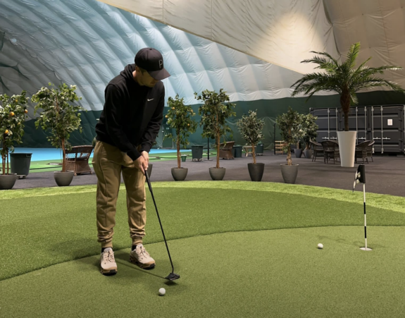 Mies mustissa vaatteissa on sisähallissa puttaamassa golf-mailalla golf-palloa. Alustana on vihreä tekonurmikko.