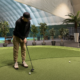 Mies mustissa vaatteissa on sisähallissa puttaamassa golf-mailalla golf-palloa. Alustana on vihreä tekonurmikko.