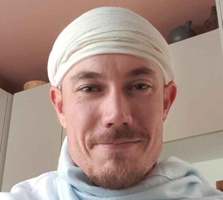Kuvassa on mies joka hymyilee, hänellä on parta ja valkoinen huivi päässä. Taustalla näkyy valkoista.