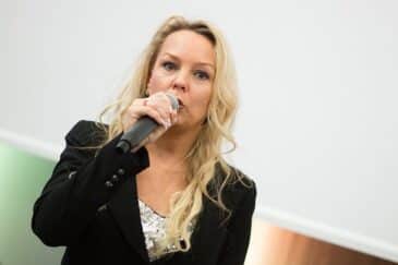 Vaaleahiuksinen nainen puhuu mikrofoniin, hänellä on musta jakku päällä. Tausta on valkoinen