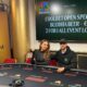 Nainen ja mies istuvat pokeripöydän ääressä, taustalla on televisio näyttö ja valkoista ja keltaista