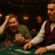 Kaksi miestä istuu pokeripöydän ääressä, toisella on vihreä paita ja toisella punainen liivi. Taustalla näkyy kaksi muuta miestä.