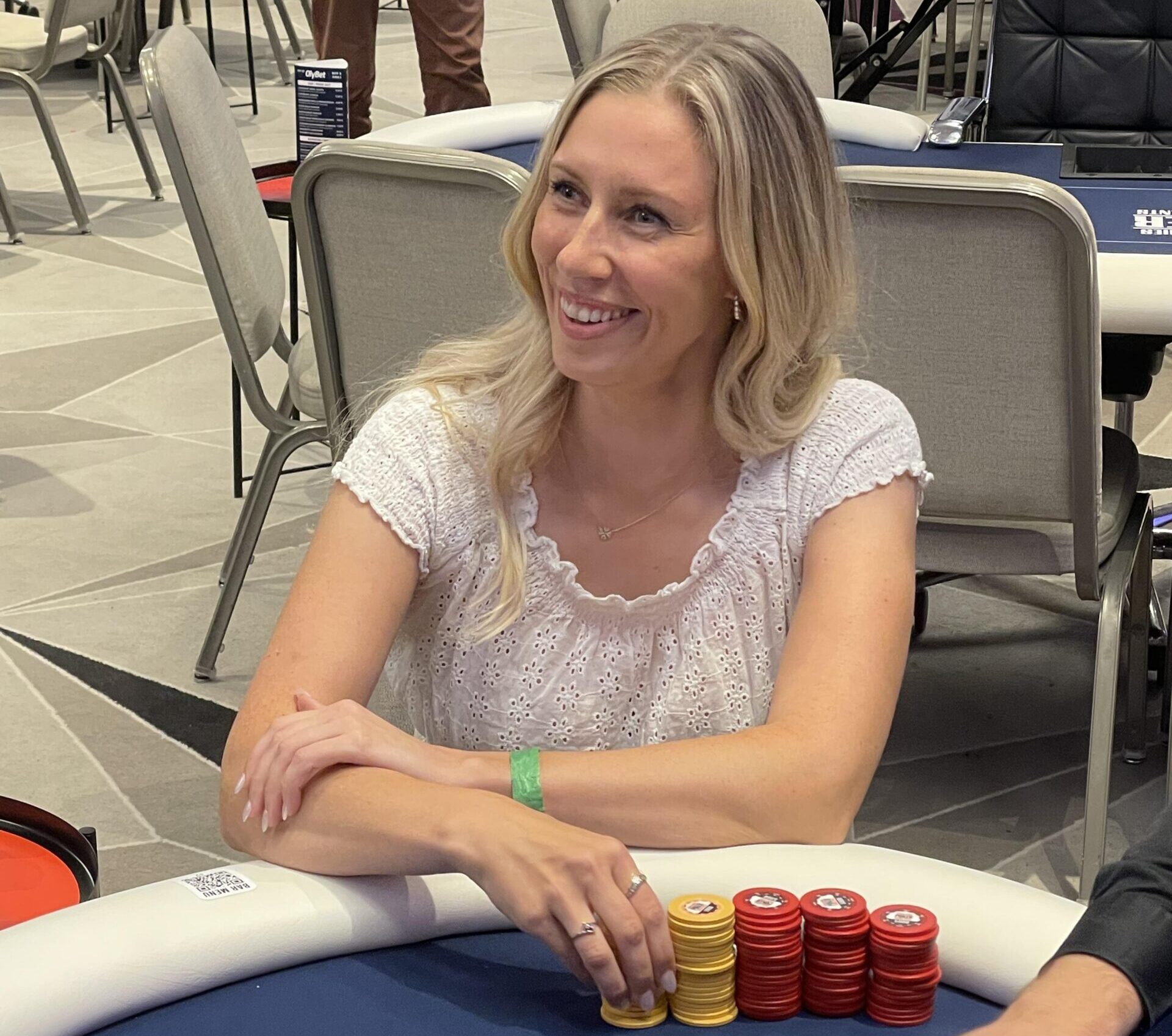 Linda Lahdenpää istuu pokeripöydän ääressä iloisesti hymyilleen valkoinen paita päällään ja kasa pokerimerkkejä edessään.