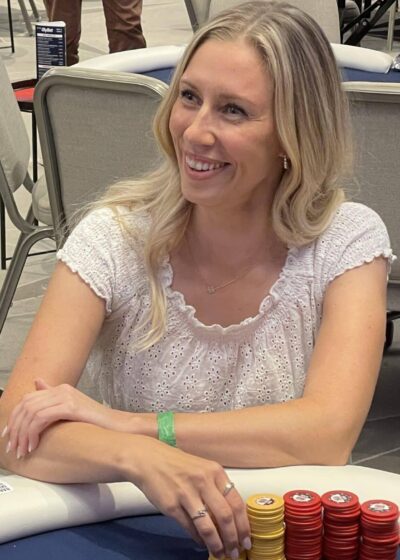 Linda Lahdenpää istuu pokeripöydän ääressä iloisesti hymyilleen valkoinen paita päällään ja kasa pokerimerkkejä edessään.